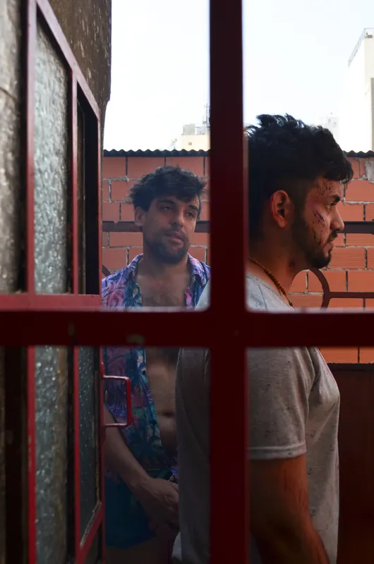 Dos personajes miran a través de una ventana de marco rojo, frente a una pared de ladrillos.