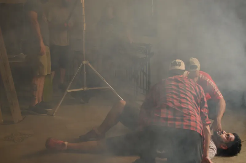 Escena con humo: dos personajes se inclinan sobre un hombre tirado en el piso, con un trípode de luz al fondo.