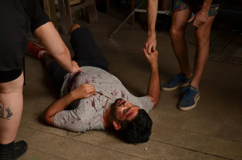 Un hombre con la remera manchada de sangre, tirado en el piso, rodeado por otros personajes.