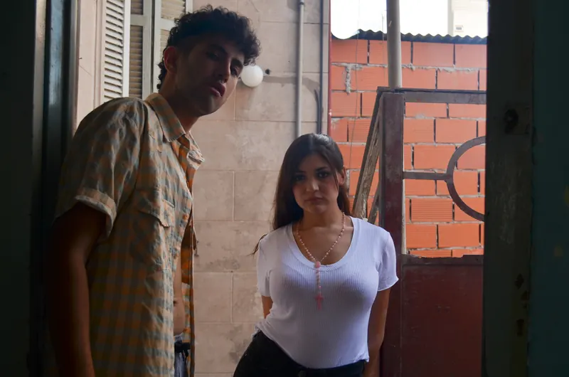 Un hombre de camisa a cuadros y una joven de remera blanca en la entrada de un patio con rejas.