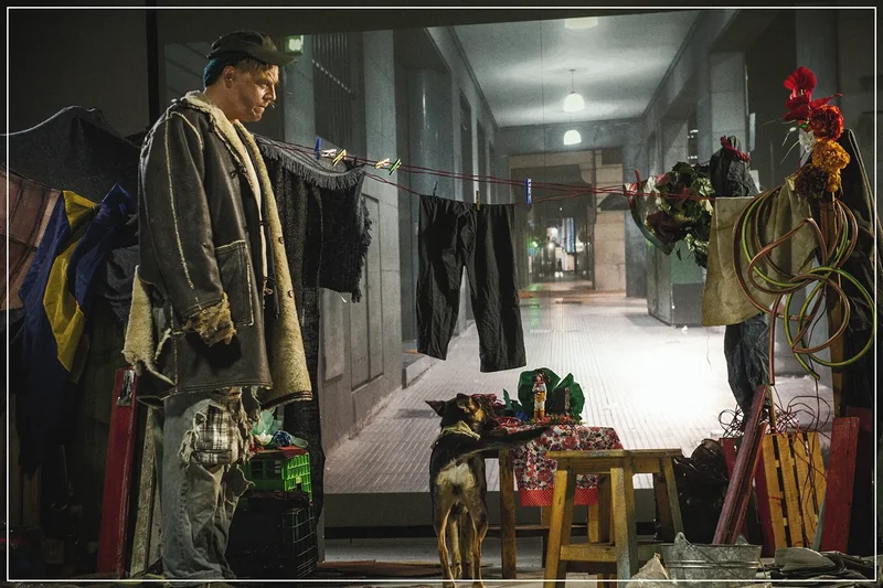 Personaje con un perro y objetos de utilería frente al fondo de un pasillo.