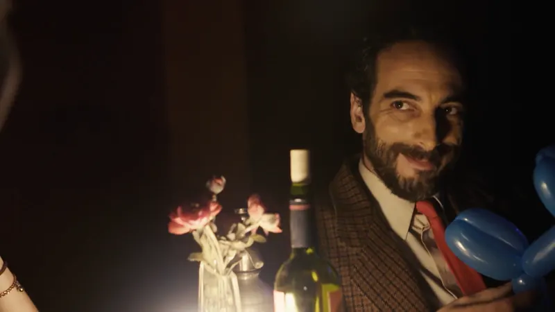 Un hombre de barba sonríe con un globo azul en la mano, junto a una mesa con flores y una botella.